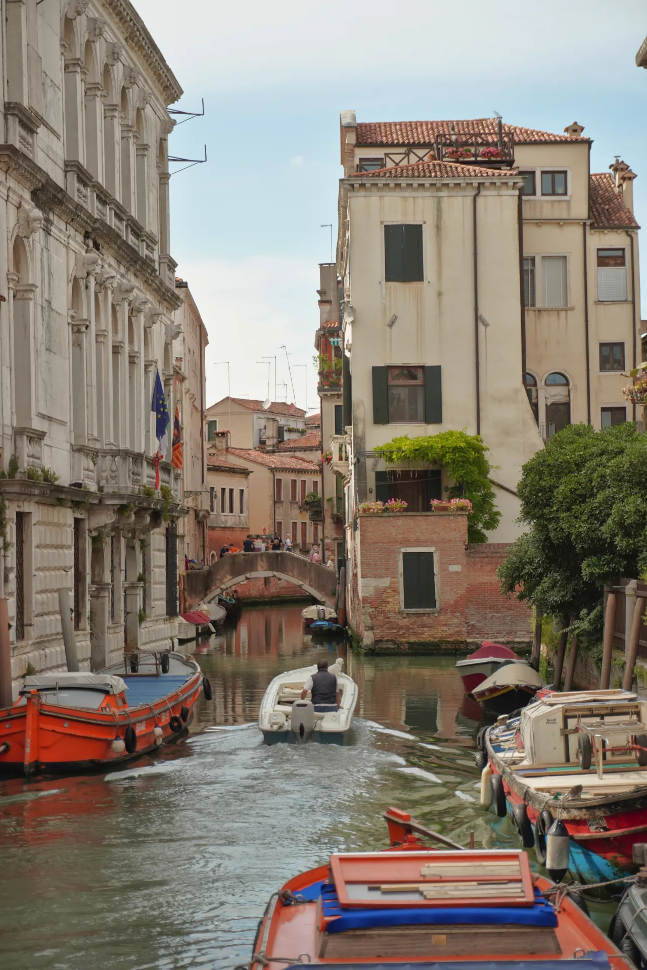 Osoba vozi mali motorni čamac niz kanal u Veneciji, sa starim zgradama sa obe strane i mostom u daljini.
