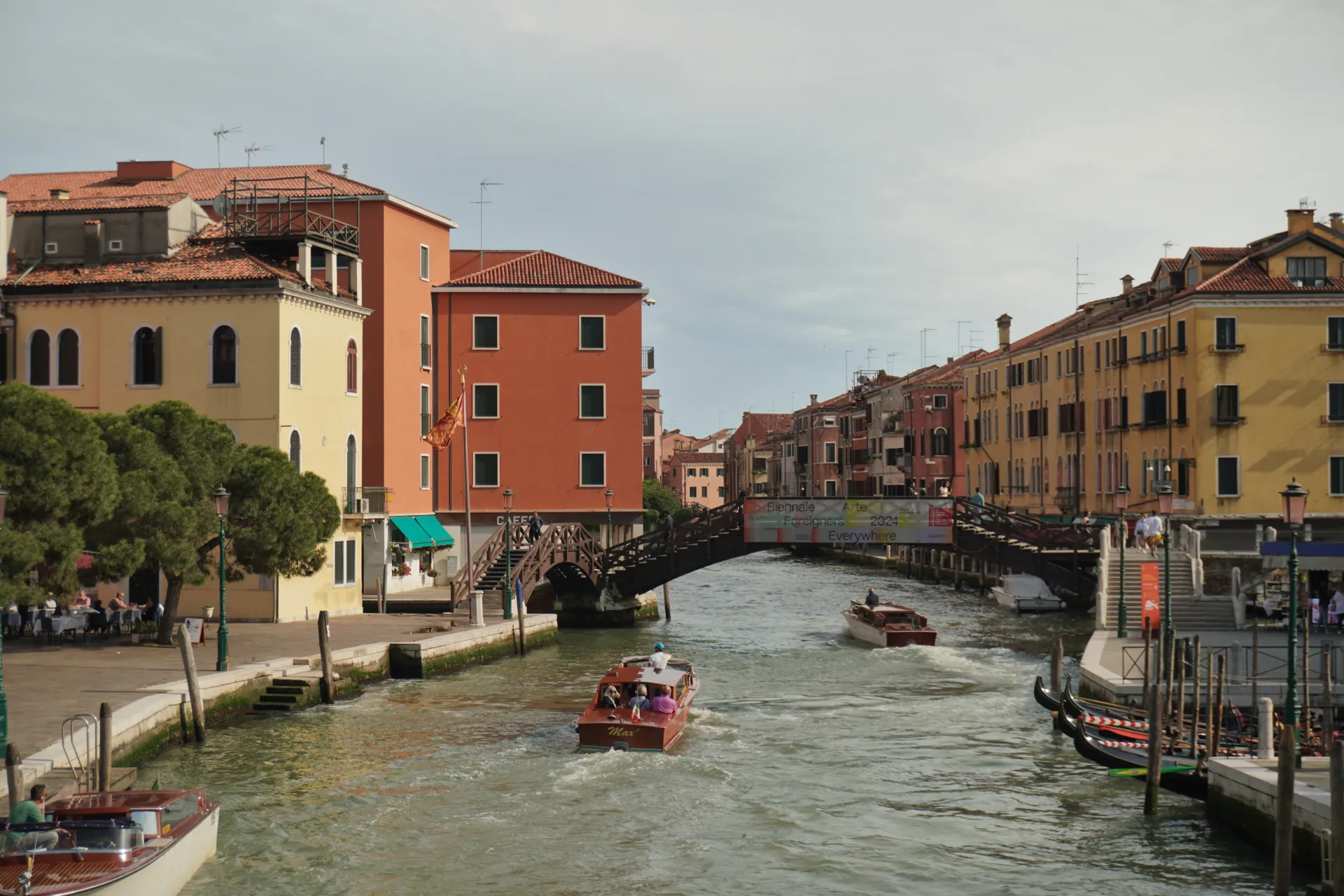 A canal in Venice, Italy, with several boats, including a water taxi and gondolas, moving through the water. A wooden bridge spans the canal, with colorful historic buildings visible on either side.