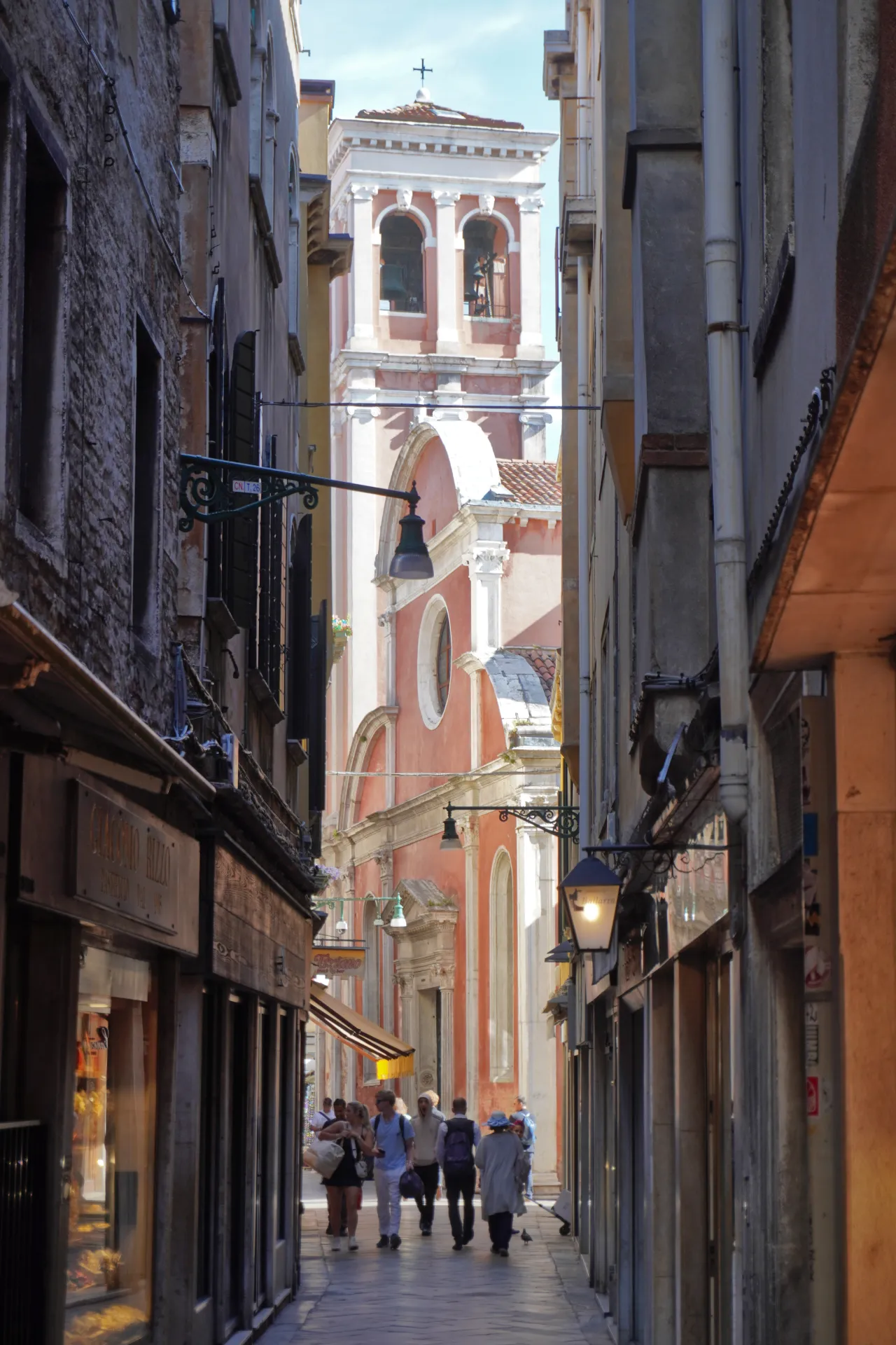 View down a narrow Venetian street towards a pink church facade and bell tower, with shops and people walking.