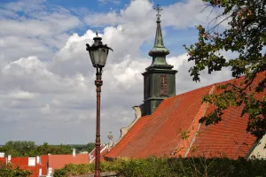 A tall, vintage street lamp with an ornate design stands to the left, with the green-spired tower of a church and a red-tiled roof visible to the right, under a sky with fluffy white clouds.
