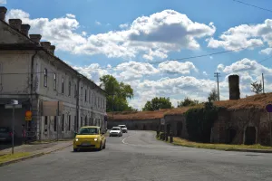 A street with a yellow car driving towards the Petrovaradin Fortress in Novi Sad, Serbia. An old building is on the left and the fortress walls are on the right.