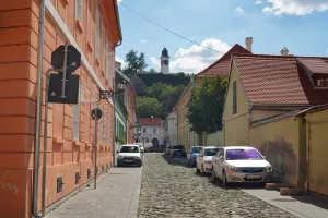 Cobblestone street in Novi Sad with colorful buildings, cars parked along the side, and the clock tower of Petrovaradin Fortress visible on a hill in the distance under a blue sky.